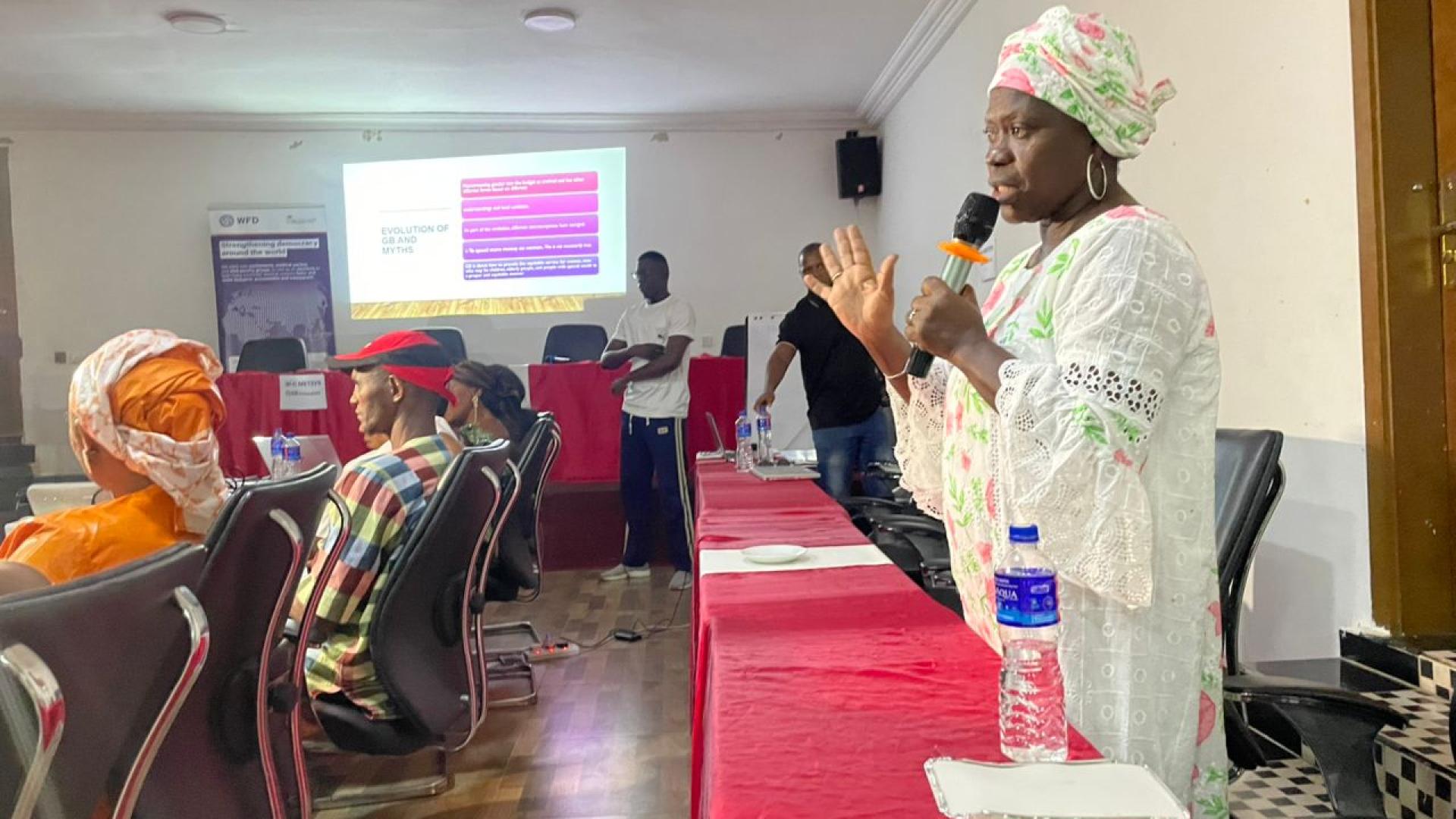 A woman speaking into a microphone, addressing workshop attendees sitting at a table