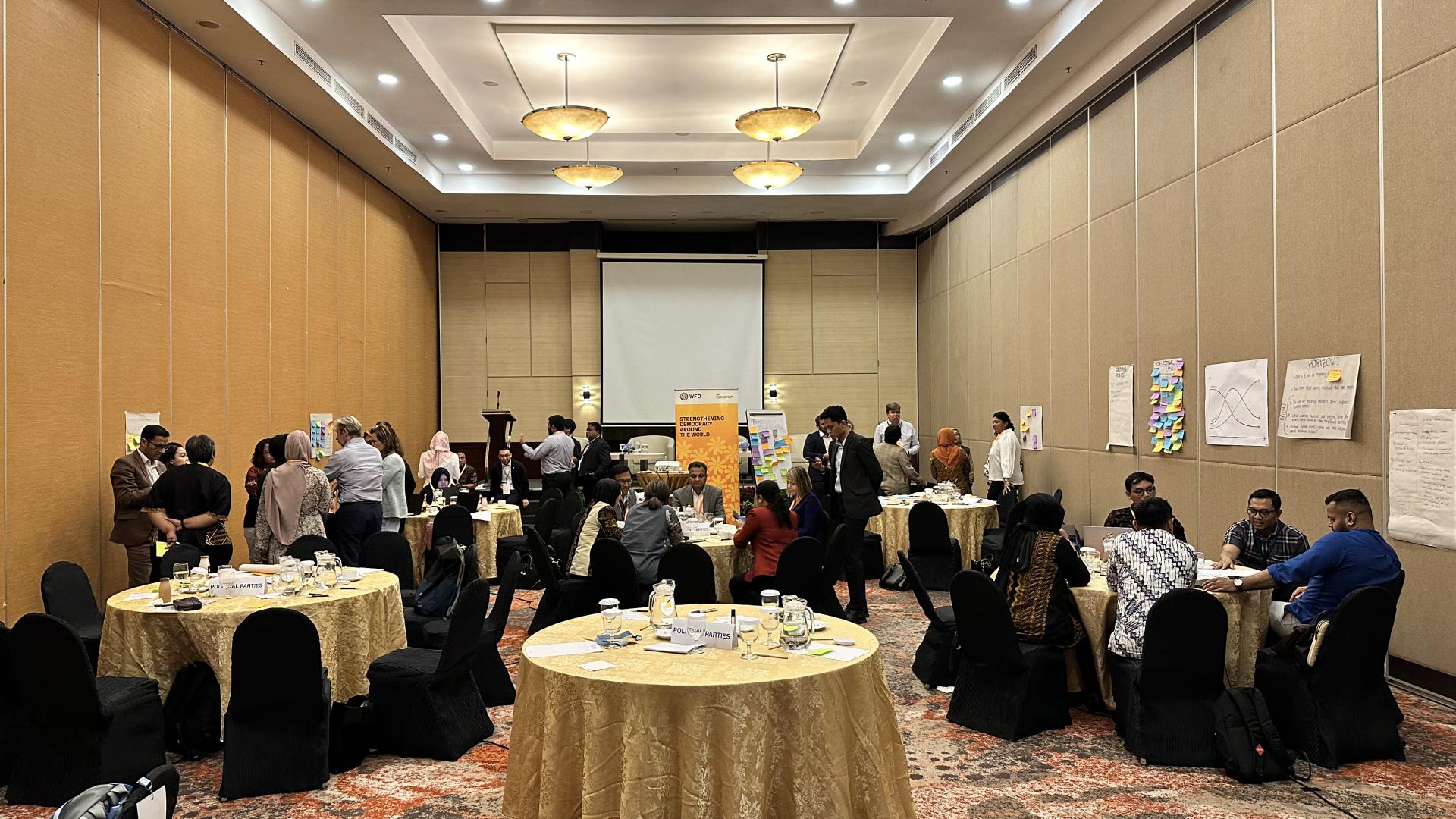 A photo of the Democracy Action Partnership event in action - a group of people are talking in groups, sat at tables or standing, concentration on brainstorming actions. In the centre is an orange banner which says strengthening democracy around the world.