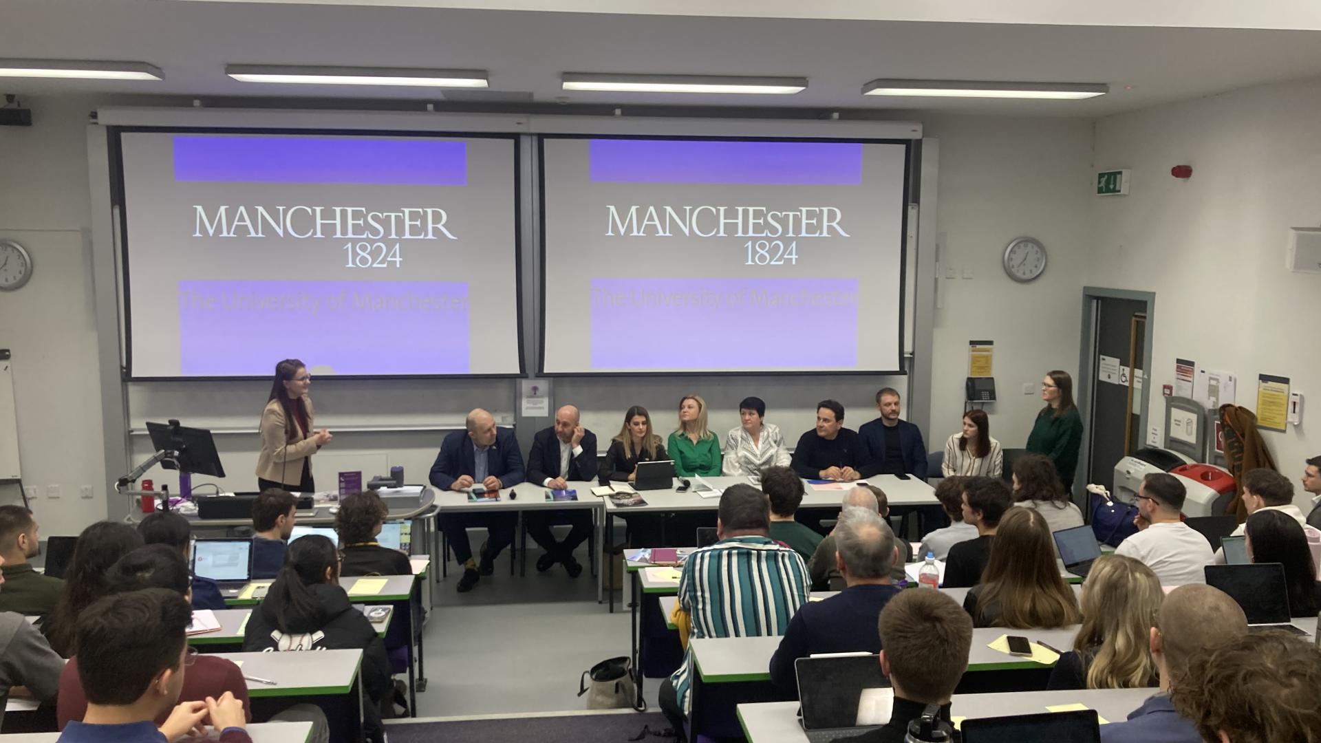 Ukrainian delegation giving a lecutre at the University of Manchester. The delegation are seating at the front of the room, and we see the backs of people's heads listening