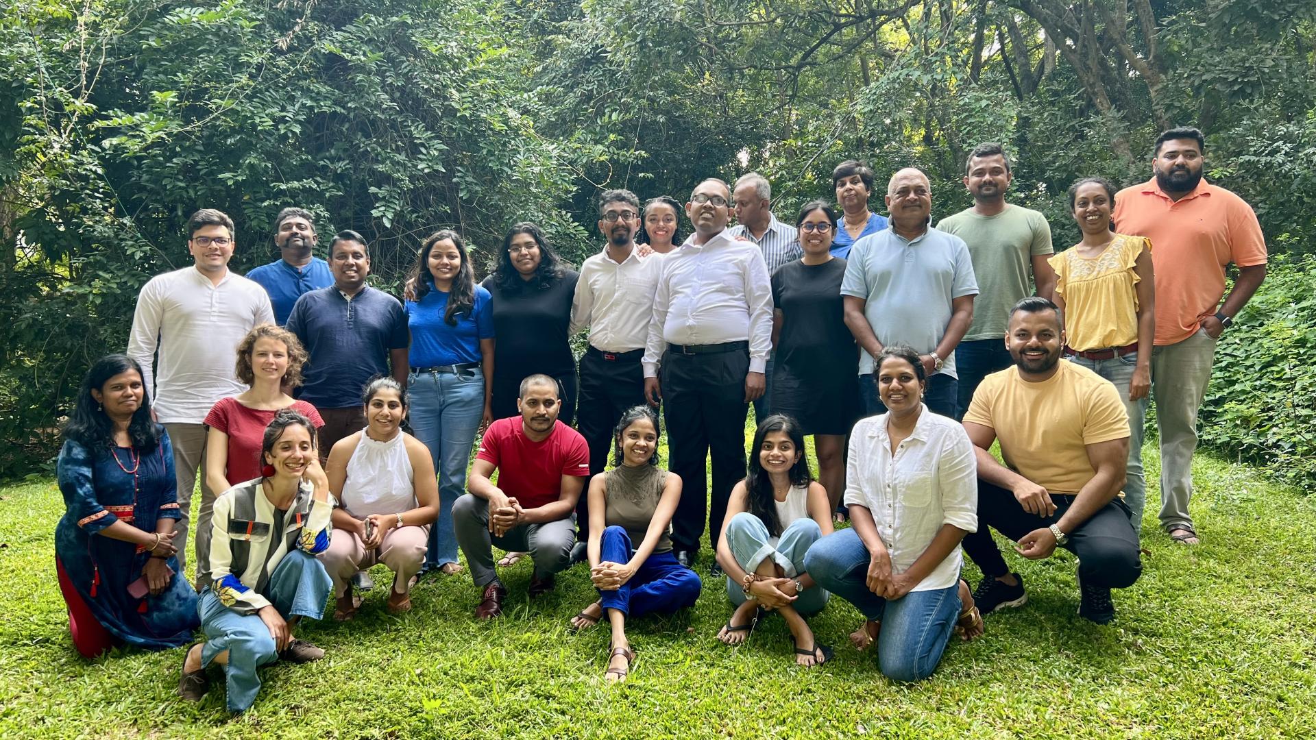 A group of people posing for a group photo. Some are seated on the grass while others are standing. There are trees seen in the background.