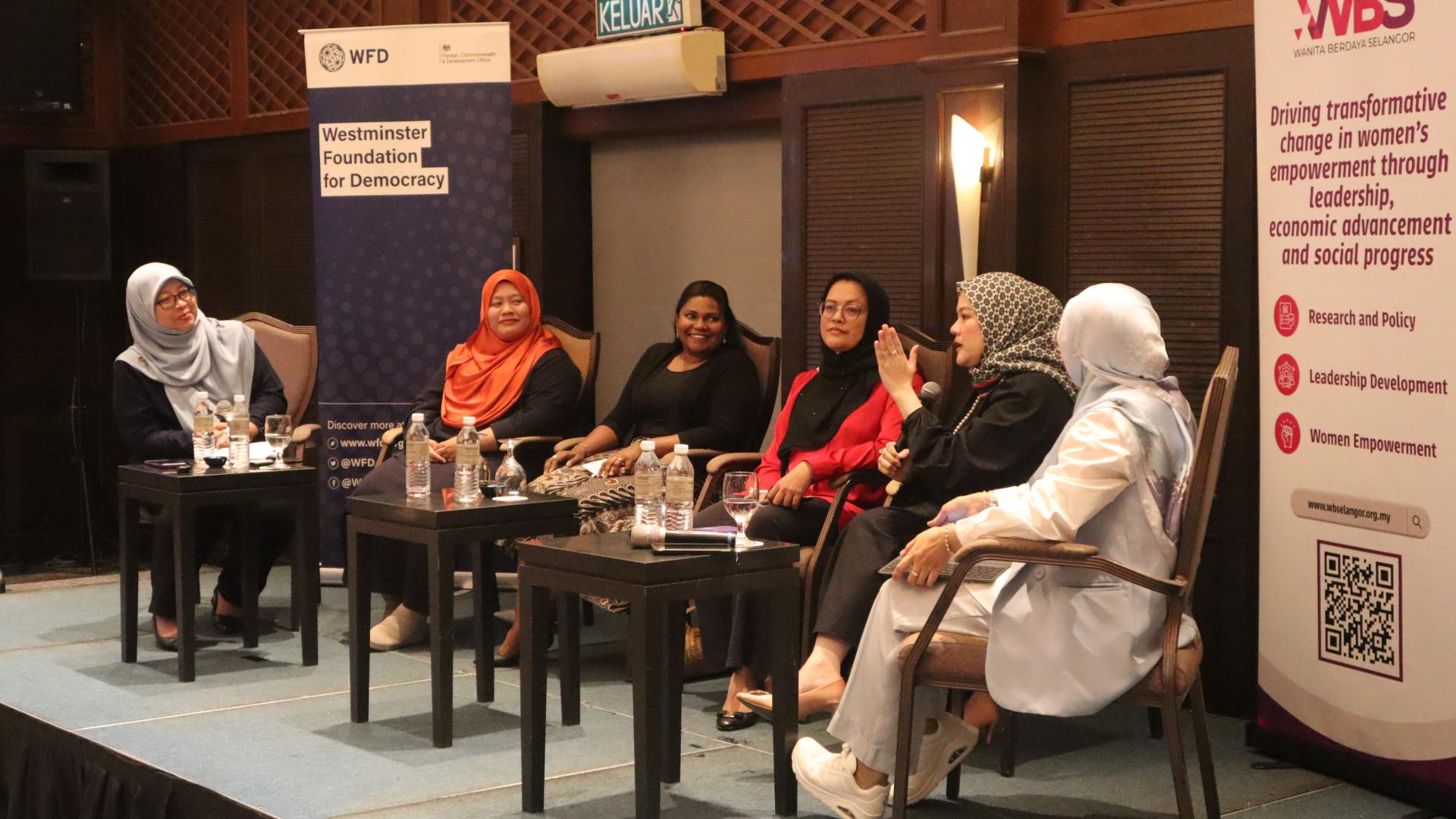 Six women are seated on a stage. One of them is speaking while others seem to be listening to her. There are side tables with water bottles in front of them. 
