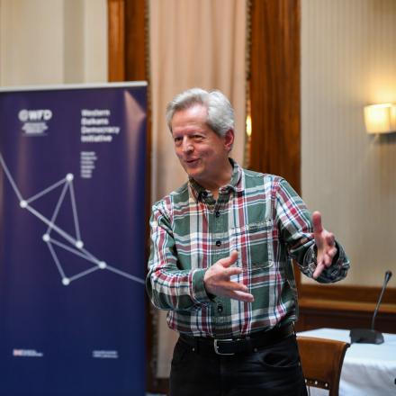 A man with grey hair speaking next to a banner for WFD's Western Balkans Democracy Initiative